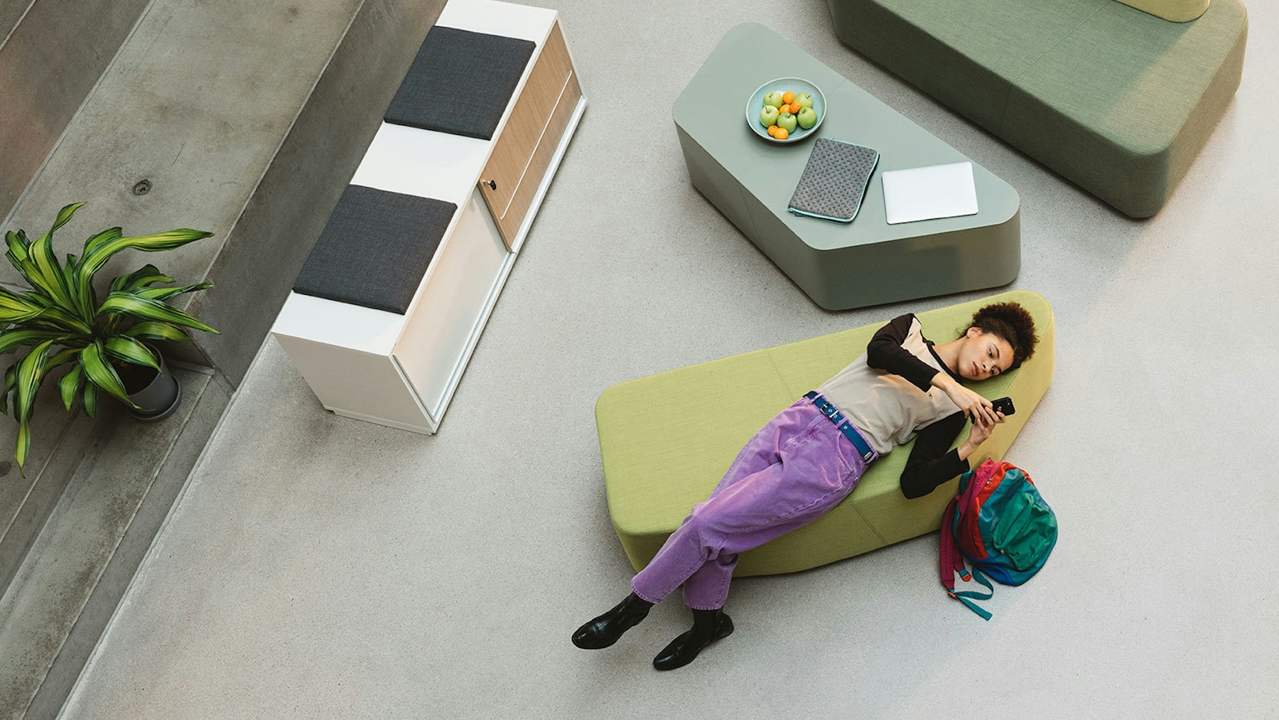 A woman with short dark hair and purple trousers is lying on an Organic Office lounge furniture, with a backpack next to her. The picture is a bird's eye view.