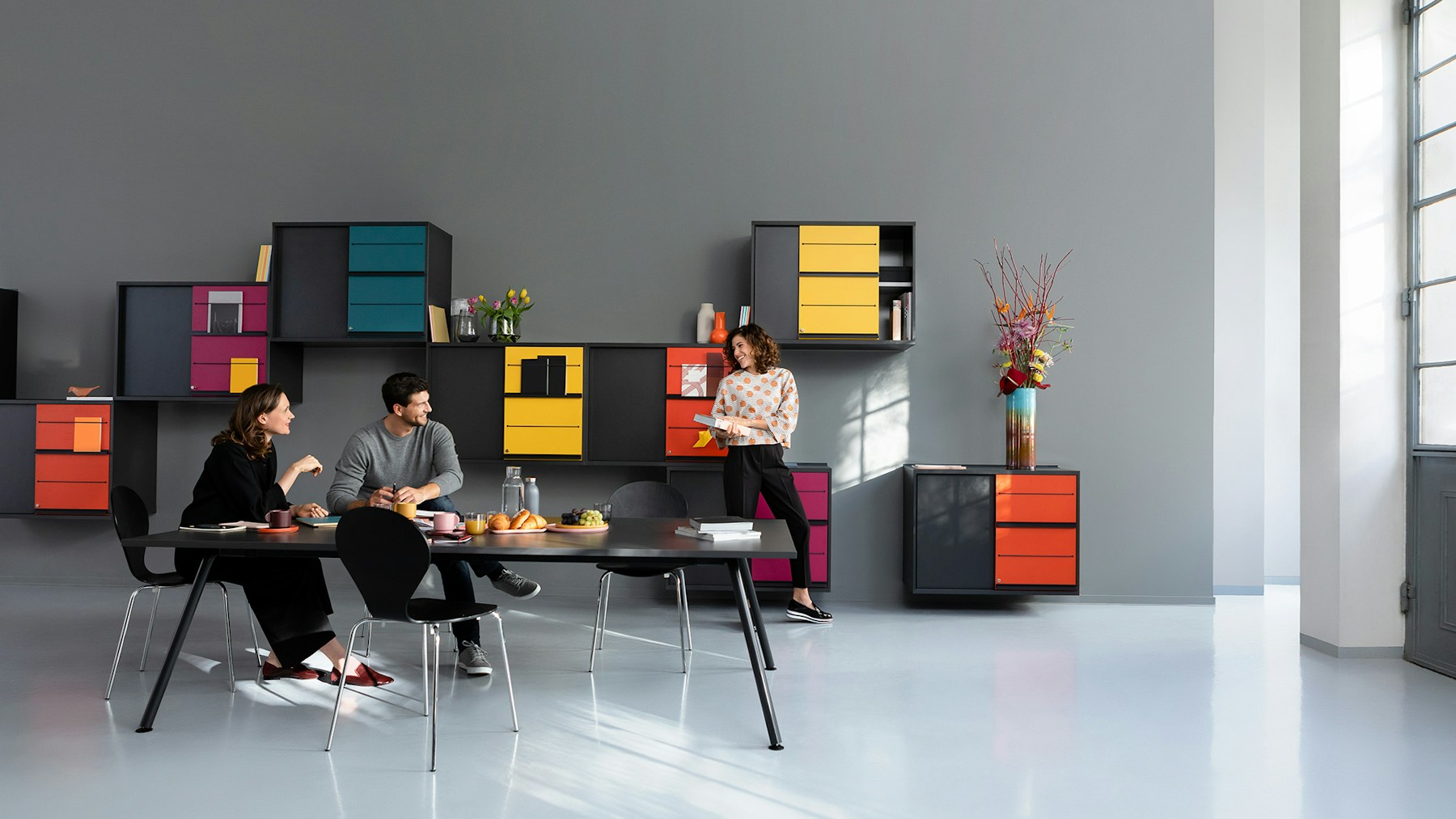 Meeting situation with a conference table in graphit and colorful cupboards on the wall. One woman and one man sit at the conference table. One woman stands in front of the cupboards.